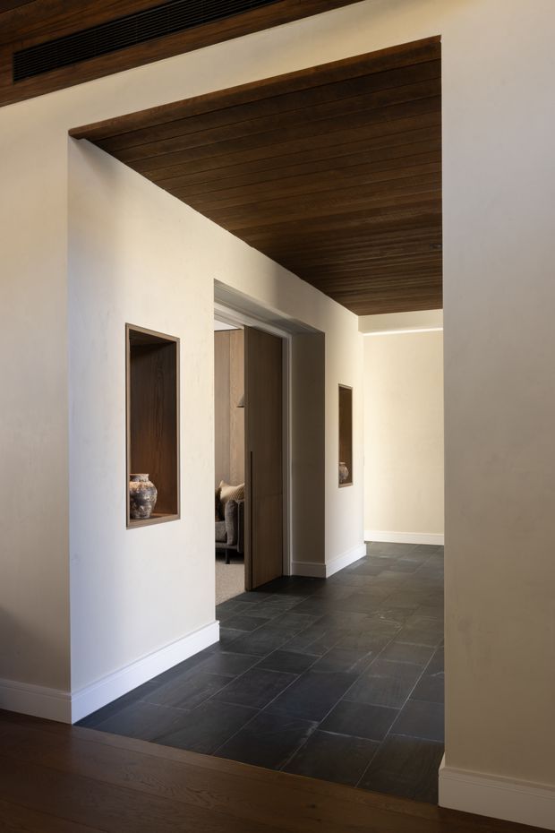 A warm, refined interior hallway or entry passage with dark slate-like tiled flooring, smooth off-white plaster walls, white baseboards, and a dramatic dark timber-lined ceiling recess. A timber door opens toward a partially visible bedroom or living area, while recessed wall niches display pottery. The space transitions to dark timber flooring in the foreground and includes a linear ceiling air vent.