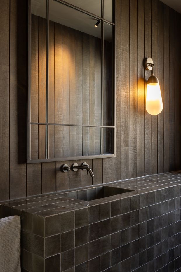 A moody, refined bathroom vanity area with dark vertically panelled walls, a large black-framed mirror, wall-mounted brushed metal tapware, and a tiled built-in basin counter. A warm sculptural wall sconce provides amber illumination and contrasts with the dark grey-brown finishes.