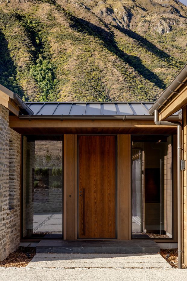 A modern residential entry courtyard viewed from outside, with a tall timber front door framed by full-height glazing, dark stone paving and steps, warm timber soffits, stone masonry walls, and a standing-seam metal roof and gutters. A steep, rugged alpine or mountain landscape rises directly behind the building.