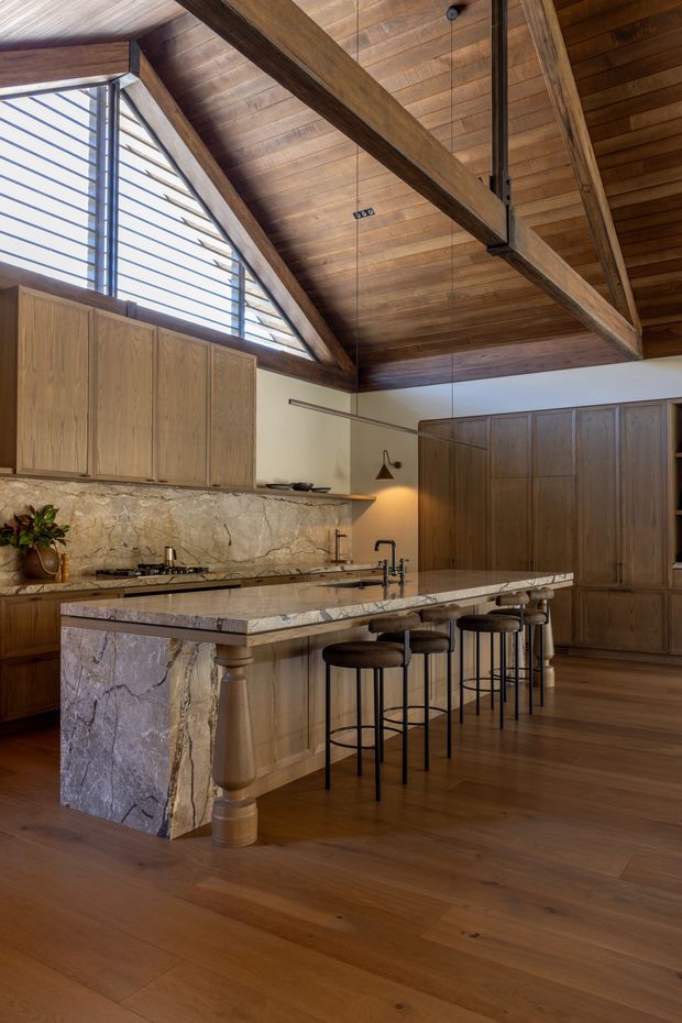 A warm, high-end kitchen with extensive timber cabinetry, a dramatic exposed timber-beam and timber-lined ceiling, and a large marble-clad island with a stone benchtop. The island includes turned timber supports and seating for several people on dark bar stools. A stone backsplash, integrated cooktop, sink, pendant lighting, and a high louvred clerestory window complete the space.