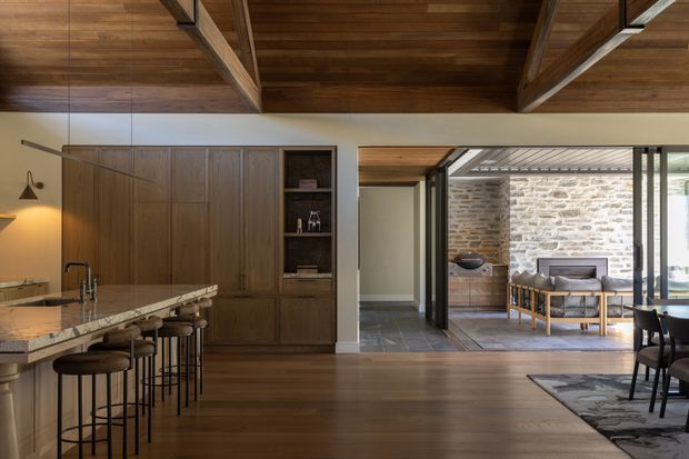 Open-plan residential interior combining a kitchen, dining area and living zone. The space features extensive dark timber cabinetry, a stone kitchen island with bar stools, timber flooring, exposed timber ceiling beams, and large glazed openings leading to a covered outdoor seating area with a natural stone wall. Warm, refined and contemporary in character.