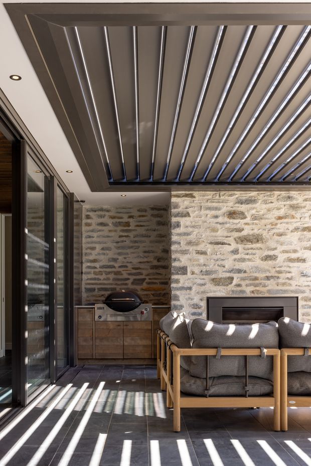 A sophisticated covered outdoor lounge or sunroom with a large operable louvred roof creating strong linear shadows. The space features exposed rough stone masonry walls, dark tiled flooring, full-height glazed sliding doors, a built-in timber cabinet with a barbecue, and substantial timber-framed upholstered lounge chairs arranged around a dark fireplace or feature opening. The design combines rustic masonry with contemporary metal and glass detailing.