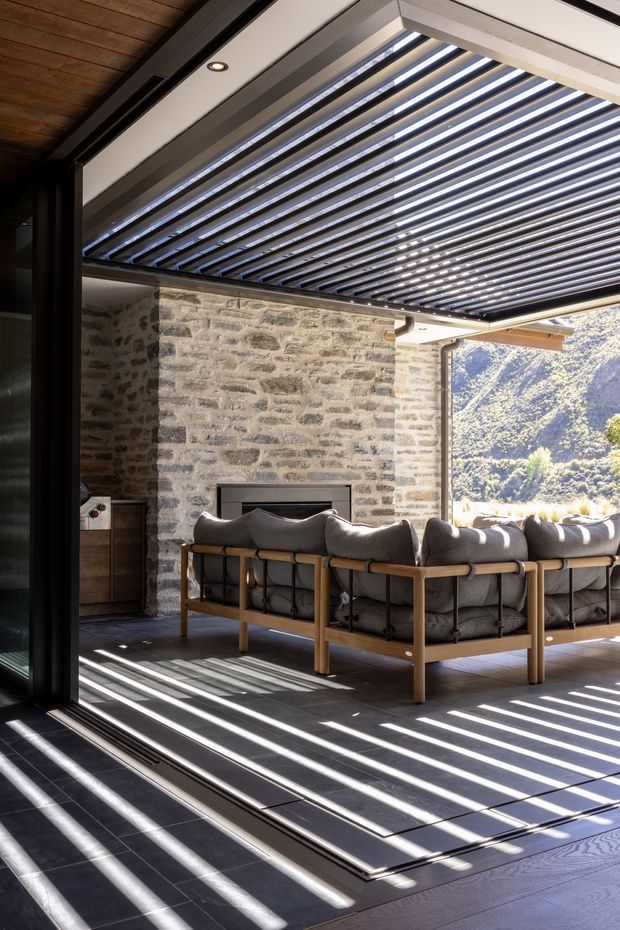 A contemporary indoor-outdoor living area viewed through large black-framed glazed doors. The space features a dark tiled floor, a timber-lined ceiling, a substantial natural stone wall with a fireplace, and a grey sectional sofa with timber framing. A louvred pergola roof casts strong linear shadows across the floor, with a sunlit mountainous landscape visible beyond.