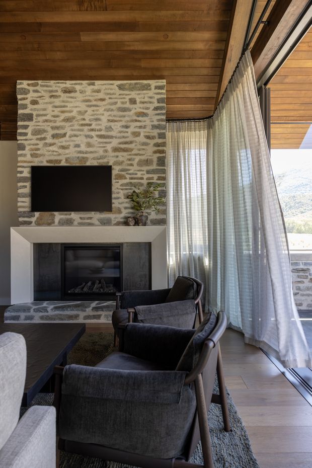 A sophisticated living room with a tall stone-clad fireplace wall, wall-mounted television, and expansive glazed corner windows dressed with sheer curtains. The room features a timber-lined ceiling, warm timber flooring, dark upholstered lounge chairs, a low dark coffee table, and a neutral textured rug. A scenic landscape is visible beyond the glazing.