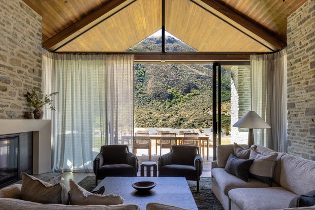A rustic contemporary living room with a dramatic timber cathedral ceiling and exposed dark beams, pale stone walls, and full-height glazed doors opening to a terrace and mountainous landscape. The room contains a large light-toned sectional sofa, two dark leather lounge chairs, a low stone or concrete coffee table, a fireplace, sheer curtains, and a sculptural floor lamp. The interior is bright, warm, and connected to the outdoors.