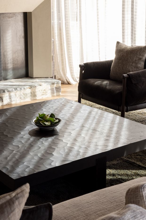 Warm, textural living room with a large sculptural metallic coffee table in the foreground, dark upholstered armchairs, a neutral sofa, sheer floor-to-ceiling curtains, a textured rug, and a minimalist concrete fireplace surround.