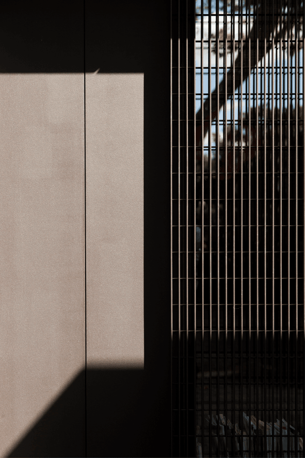 Abstract close-up architectural facade detail featuring a dark matte wall or panel beside a tall grid of narrow vertical metal screens or grilles. Strong sunlight creates sharply defined geometric shadows across the pale wall, while reflective glazing and structural elements are visible behind the grille. The composition is minimalist, high-contrast, and predominantly monochromatic.