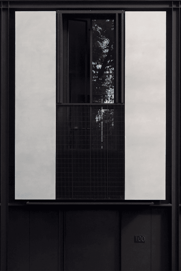 A dark, minimalist multi-storey building facade viewed straight on. The composition features large smooth white vertical panels framing a recessed central opening with black-framed glazing, a finely gridded dark metal screen or balustrade, and dark vertical cladding below. A small plaque marked “100” is visible near the lower right. The facade has a restrained monochrome palette and strong geometric symmetry.