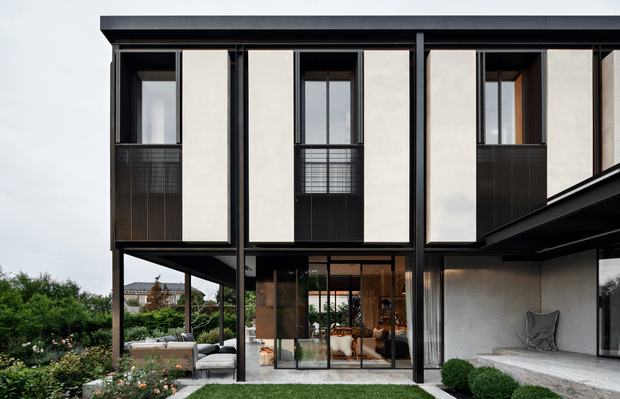 Contemporary multi-level residential facade with a strong rectilinear composition, flat roof, slim dark structural framing, pale rendered wall panels, and tall glazed openings. The elevated upper storey projects over a landscaped outdoor living area, with balconies screened by dark metal mesh and a glazed ground-floor connection to the interior. Black-framed glazing, a covered terrace, clipped shrubs, and garden planting reinforce the refined modern design.