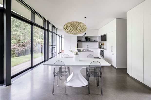 Bright open-plan kitchen and dining area with extensive black-framed floor-to-ceiling glazing overlooking a garden. The space features a large white dining table with transparent acrylic chairs, minimalist white full-height cabinetry, a compact kitchen with island, polished grey concrete flooring, and a woven pendant light. The overall design is clean, contemporary, and light-filled.