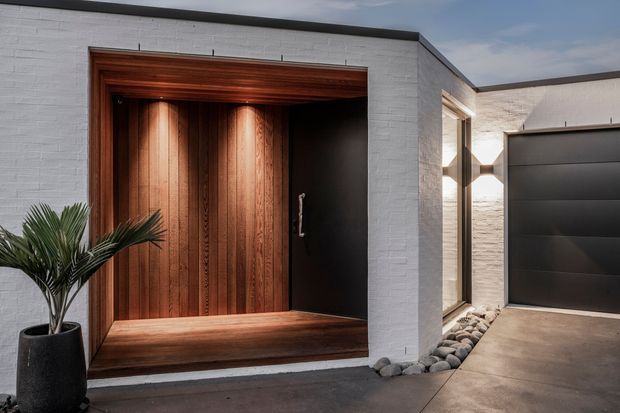 Contemporary residential entry facade featuring a deep timber-lined recessed doorway with a broad timber threshold and black door, set into a white textured masonry wall. A matte dark garage door and narrow glazed opening are visible to the right, with a pebble drainage strip and potted palm completing the composition.