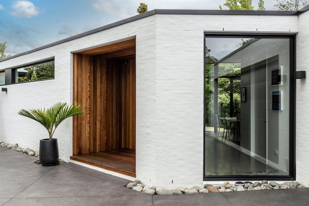 A contemporary single-storey home facade with a flat roof, white textured masonry walls, dark-framed floor-to-ceiling glazing, and a deeply recessed timber-clad entry. A smooth concrete forecourt and decorative river stones line the base, with a potted palm beside the entrance. Interior furnishings are faintly visible through the glazed doorway.