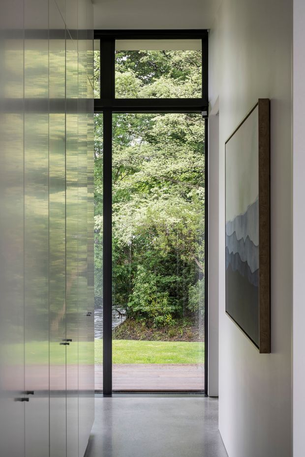 A narrow, minimalist interior passage beside a full-height glazed door and tall black-framed windows opening to a leafy garden. The space features smooth white walls, a polished grey concrete floor, reflective full-height built-in cabinetry or glazing on the left, and a large framed artwork on the right. Timber decking and dense greenery are visible outside.