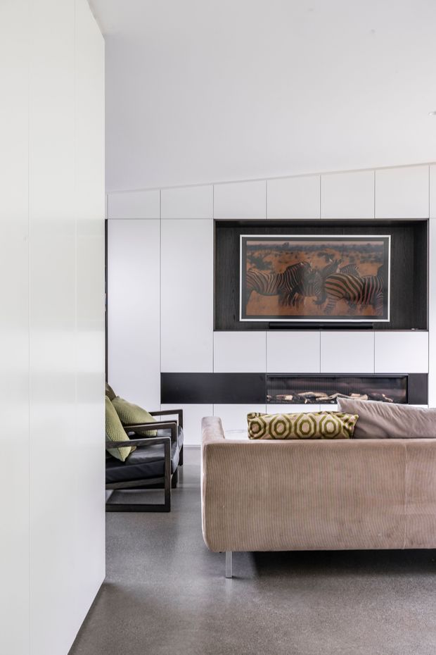 A contemporary living room viewed from behind a partial white wall. The room features a large pale upholstered sofa, black-framed armchairs with green cushions, grey carpet, and a full-height white built-in media wall. A dark recessed display niche contains framed artwork, with a black horizontal fireplace or media element below. The scheme is restrained and monochromatic with olive-green and warm neutral accents.
