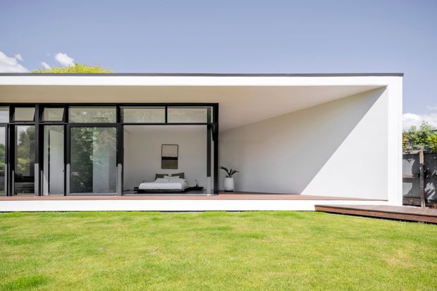A minimalist modern single-storey house exterior with a long flat roof and a deep covered terrace. Black-framed floor-to-ceiling glazing opens onto a bedroom visible inside, while a sculptural white angled wall forms a sheltered outdoor alcove. A timber-toned deck borders the manicured lawn.