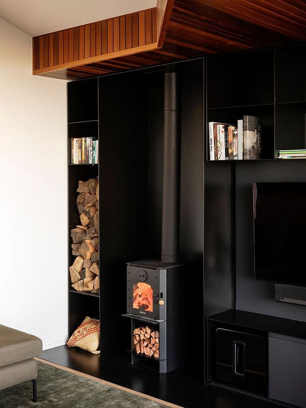 A contemporary living room media wall featuring a black built-in shelving and cabinetry system, an enclosed wood-burning stove with a vertical flue, firewood storage, bookshelves and television. Warm timber slatted ceiling and bulkhead detailing contrast with the dark joinery, while a sectional sofa and rug are partially visible.