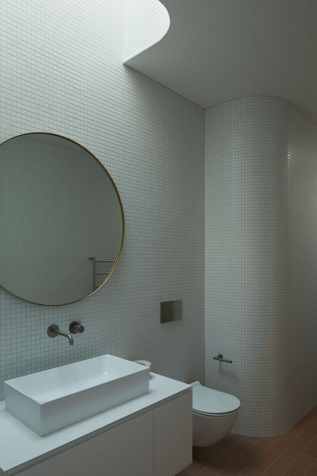 A compact contemporary bathroom with pale small-format mosaic wall tiles wrapping around curved walls and a rounded shower enclosure. The room features a large circular mirror with a thin metallic rim, a wall-mounted basin tap, a white rectangular vessel basin on a streamlined vanity, concealed toilet cistern, and warm brown floor tiles. The curved tiled surfaces and restrained fittings create a refined, minimalist appearance.