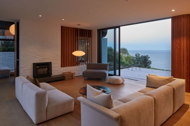 A refined contemporary living room with a large sectional sofa arranged around a low coffee table and a built-in fireplace set into a textured white masonry wall. Full-height black-framed glazing opens to a timber deck with expansive ocean views. Vertical timber screening and warm pendant lighting add texture and warmth to the otherwise neutral interior.