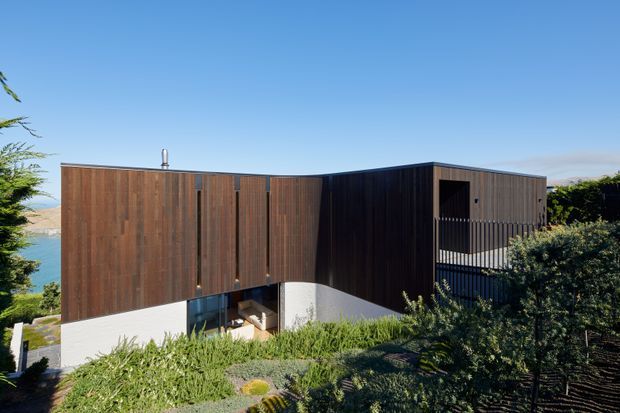 Exterior view of a contemporary multi-level home on a sloped, vegetated site. The building has a strongly articulated dark timber-clad upper volume, lighter rendered lower walls, a flat roof, deep recessed openings, and a projecting balcony or terrace. Water is visible beyond the left side of the property.