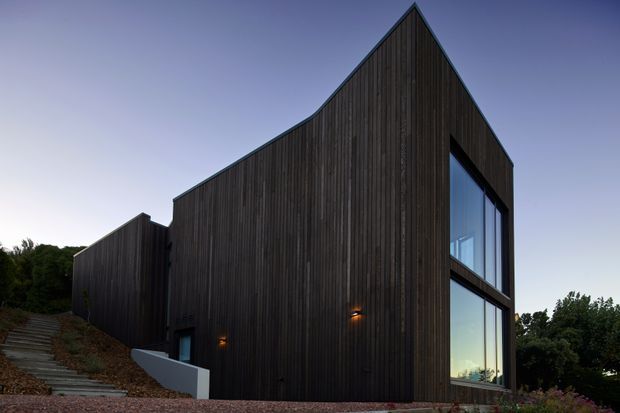 A dark, sharply angular contemporary house exterior with tall vertical timber cladding, large stacked corner windows, and a steep asymmetrical gabled roofline. The building sits on a sloping, landscaped site with an exterior stairway and subtle wall lighting, photographed at dusk.