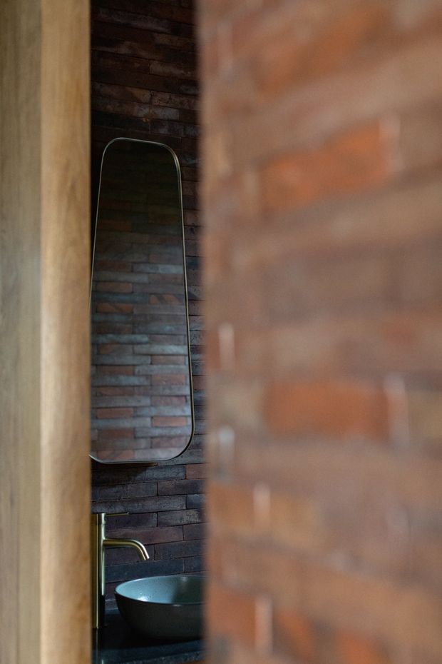 A narrow, moody bathroom view framed through a doorway or partial brick wall. The space features dark exposed brick wall finishes, a rounded rectangular mirror, a black vessel basin on a dark vanity surface, and a brushed metal wall-mounted tap. Warm timber framing contrasts with the deep red-brown brickwork.