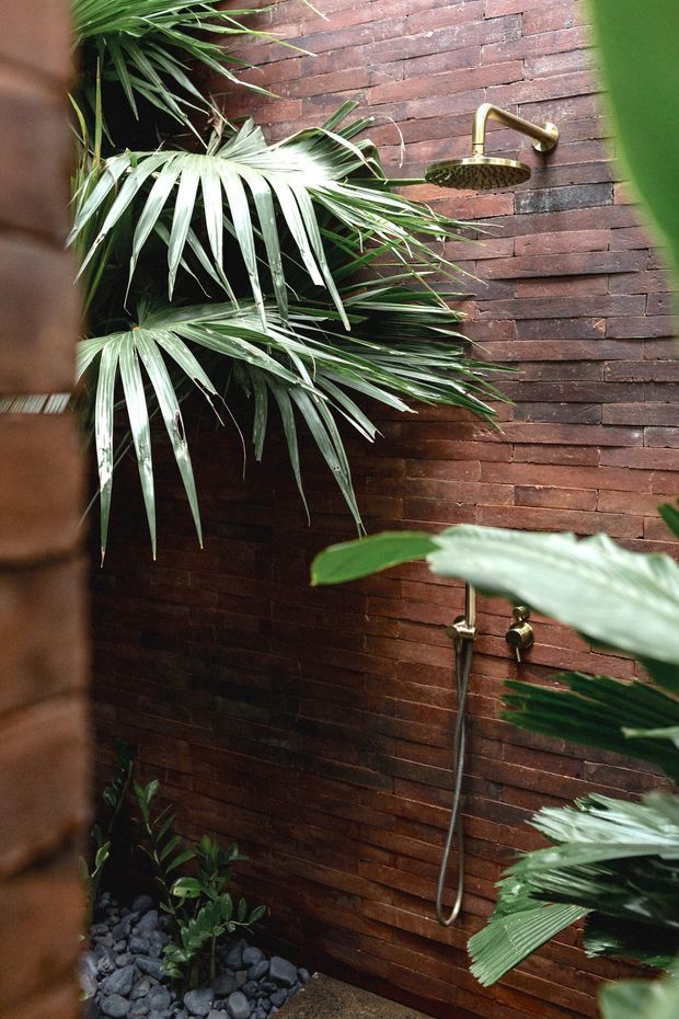 A tropical outdoor shower set against a dark reddish-brown brick wall. The wall-mounted brass rain shower and handheld shower are surrounded by lush palm foliage, with a small black-stone garden bed at the base. The composition is intimate, natural and spa-like.