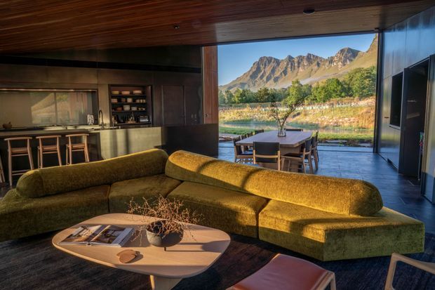 A warm, open-plan contemporary living room with a large olive-green sectional sofa and sculptural wood coffee table in the foreground. Dark timber ceiling, cabinetry and wall finishes frame a compact kitchen at left, while full-height glazing opens to an outdoor dining terrace and a dramatic mountain landscape beyond. The interior uses rich natural materials and strong indoor-outdoor connection.