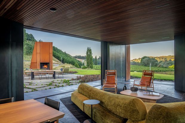 A refined open-plan living and dining space with full-height glazing opening onto a landscaped outdoor terrace and garden. The room features a dark timber slatted ceiling, dark metal structural framing and wall surfaces, a sculptural ochre-green curved sofa, mid-century-style lounge chairs, and a timber dining table. Outside, a large built-in fireplace and paved terrace are clearly visible, with rolling greenery beyond.