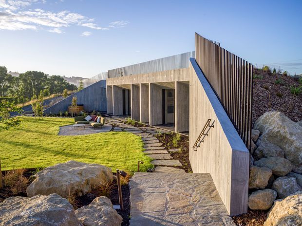 A low-profile contemporary concrete building integrated into a landscaped, rocky sloping site. The facade features a series of recessed openings beneath a flat concrete roof, with a tall vertical timber screen rising along one side. A paved stepping path leads through a manicured lawn toward the building, with large natural boulders forming the foreground and retaining landscape.