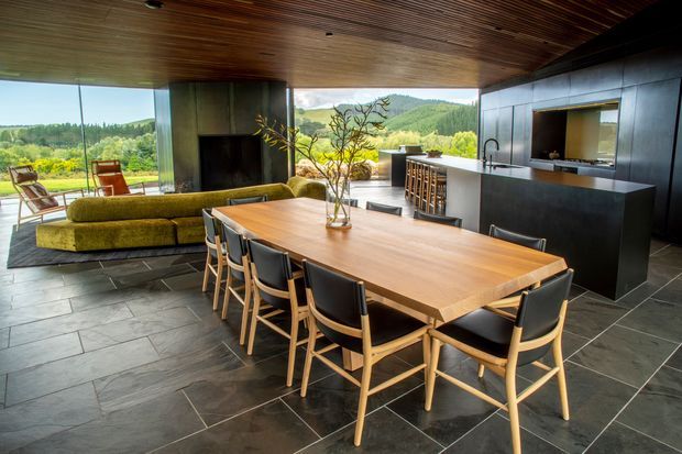 Open-plan contemporary kitchen, dining and living area with a large timber dining table, black-framed dining chairs, a dark kitchen island and cabinetry, slate-like tiled flooring, and a timber-lined ceiling. Floor-to-ceiling glazing opens to a green hilly landscape, while a fireplace and low sofa define the living zone.