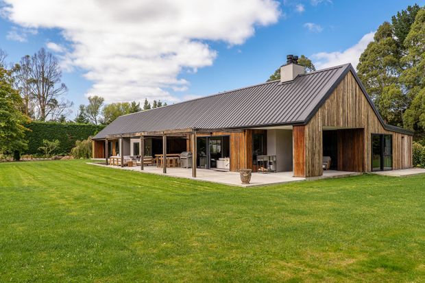 A single-storey rural-style residence with a long pitched gable roof, dark vertical metal roofing, extensive timber cladding and large glazed openings. A covered outdoor veranda with timber posts and outdoor seating runs along the front, opening onto a broad manicured lawn and landscaped garden.