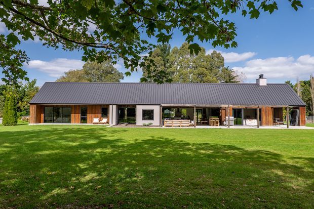 A wide exterior view of a long, low-slung contemporary home set behind an expansive lawn. The house has a dark grey pitched metal roof, extensive floor-to-ceiling glazing, warm timber cladding and framed openings, with a covered central outdoor living area and landscaped garden surrounding it.