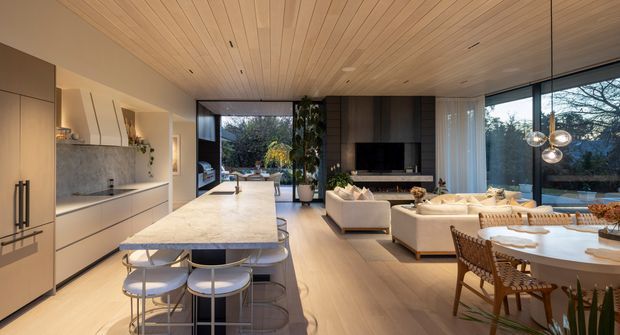A bright, open-plan kitchen, dining and living space with a large marble island and integrated light cabinetry in the foreground. The room features a warm timber-lined ceiling, pale timber flooring, floor-to-ceiling glazing, a contemporary fireplace wall, sculptural pendant lighting, and direct views to an outdoor terrace and garden.
