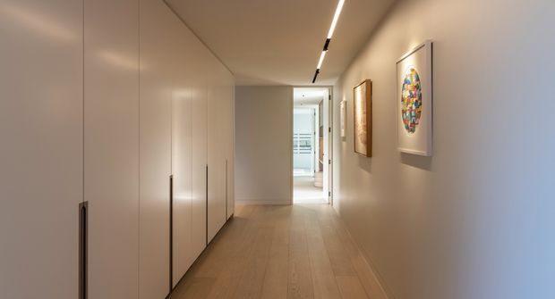 A long, narrow contemporary hallway with warm timber flooring, full-height minimalist built-in cabinetry along the left wall, smooth light-coloured walls, recessed linear ceiling lighting, and framed artwork on the right. A brighter room is visible at the far end.