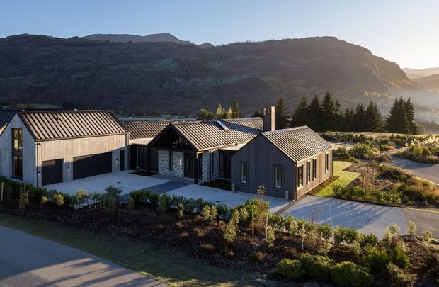 A contemporary single-storey residential compound set against a dramatic rocky mountain landscape. The dark charcoal and grey buildings have steep pitched gable roofs with standing-seam metal roofing, vertical dark cladding, pale stone masonry, and extensive garage and entry glazing. Landscaped planting, paved access areas, and tall conifers surround the house.