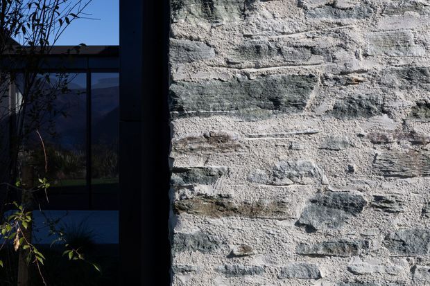 Close-up architectural view of a rough, light grey natural stone masonry wall beside dark-framed glazing. The glazing reflects or reveals a dim landscaped exterior with silhouetted branches and distant terrain, while the stonework dominates the composition. The image has a high-contrast, moody atmosphere.