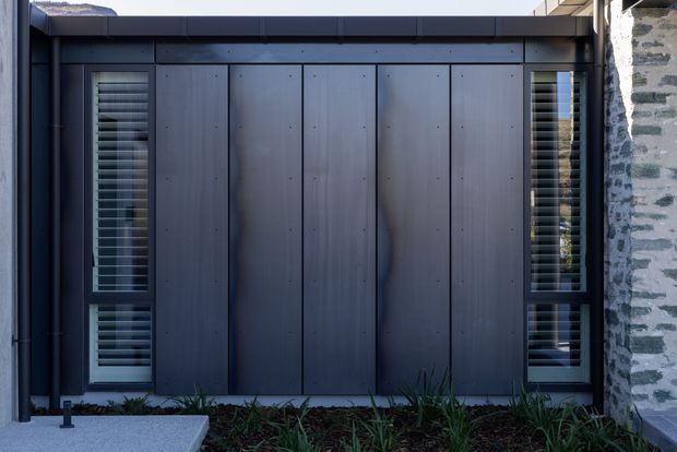 Close-up exterior view of a contemporary facade clad in dark vertical panels, with narrow windows fitted with horizontal louvres on either side. A dark projecting roof or parapet element caps the facade, while a contrasting rough grey masonry wall is visible at the right edge. Low planting borders the base.