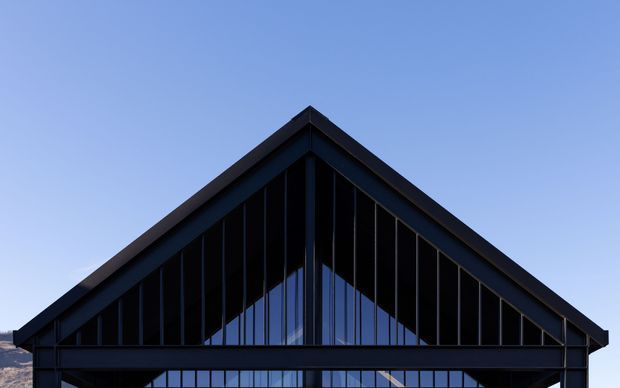 Close-up exterior view of a contemporary dark-clad gable-front building. The sharply pitched triangular roofline frames a glazed gable with strong vertical mullions and exposed structural framing, set against a clear blue sky.