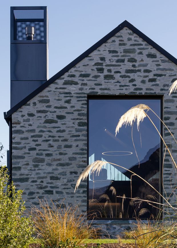 Close-up exterior view of a contemporary gable-front building clad in irregular grey stone masonry, with dark metal roof edging, a tall dark metal chimney or flue structure, and a large reflective glazed opening. Ornamental grasses and planting frame the facade, while the glazing reflects surrounding mountain scenery and pendant-like interior lights.
