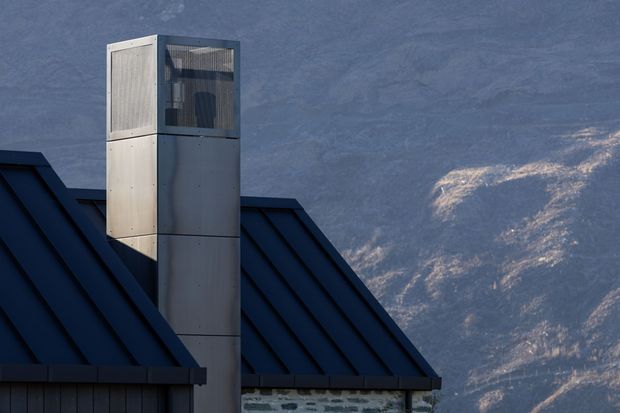 Close-up exterior architectural detail showing a dark standing-seam metal pitched roof with a tall modern metal-clad chimney or flue enclosure. The chimney has a perforated upper section and smooth grey metal panels below. A rugged mountain landscape is visible in the background.