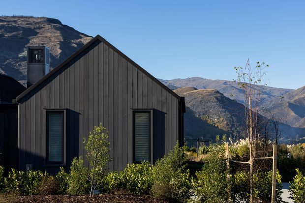Exterior view of a compact dark-clad gable-roof building set within landscaped planting, with a dramatic mountainous backdrop. The facade features vertically oriented dark timber-like cladding, simple black-framed windows with horizontal blinds, and a projecting chimney or flue element.
