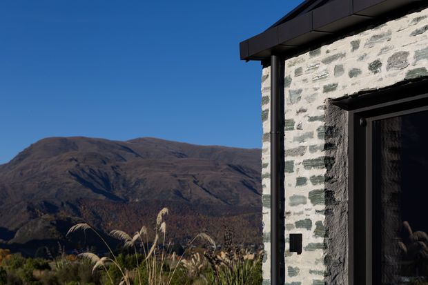 Exterior architectural detail of a contemporary mountain dwelling, showing a pale rendered masonry wall with exposed stone-like texture, dark metal roof edging, dark vertical downpipe, and a black-framed glazed doorway. A rugged mountain landscape is visible beyond the building, with ornamental grasses in the foreground under clear blue sky.