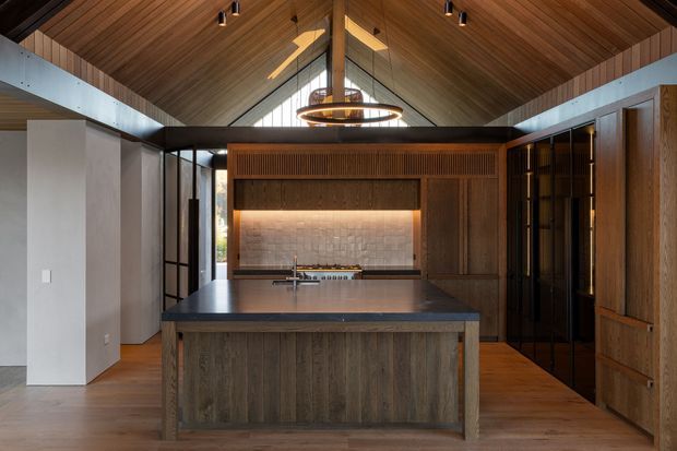 A refined contemporary kitchen with a large central island, dark timber cabinetry, and a dramatic pitched timber-lined ceiling. The island has a dark stone benchtop and integrated sink, while warm under-cabinet lighting highlights a textured stone backsplash. Black-framed glazing and a sculptural circular pendant add contrast, with abundant timber creating a warm, sophisticated atmosphere.