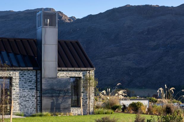 Exterior view of a contemporary mountain house with pale rough stone walls, a steep dark metal roof, and a tall metal-clad chimney or tower. The building sits in a landscaped garden with ornamental grasses, backed by a dramatic rugged mountain range under a clear blue sky.
