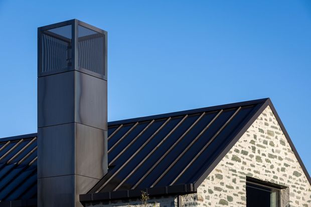 Close-up exterior view of a contemporary pitched-roof building featuring a dark standing-seam metal roof, a tall square metal chimney or flue enclosure, and a pale irregular stone masonry gable wall with dark-framed openings. Clear blue sky forms the background.