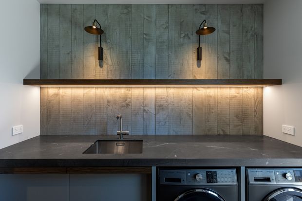 Contemporary laundry room with a dark stone benchtop, integrated sink and tap, front-loading washing machines, and extensive vertical grey timber wall panelling. Warm under-shelf lighting and two bronze wall sconces illuminate the work area.