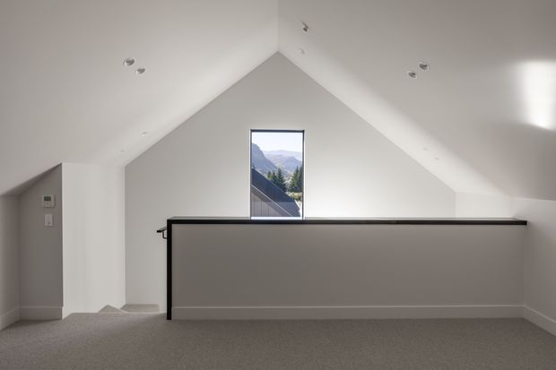 A bright, minimalist upper-level landing or loft space beneath a steep pitched roof. White walls and sloping ceiling planes frame a narrow vertical window with mountain views. A simple dark-framed half-height balustrade overlooks the level below, and the floor is finished in neutral grey carpet. Recessed ceiling lights and clean white skirting create a restrained contemporary interior.