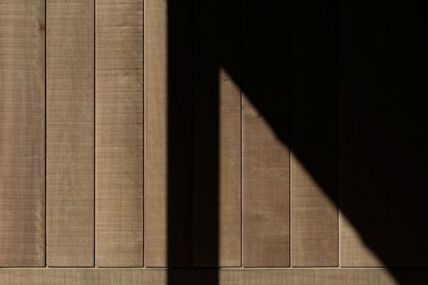 Abstract architectural detail showing vertically aligned warm brown timber wall or cladding boards with narrow dark gaps. Strong directional sunlight creates a dramatic triangular shadow across the textured wood surface, emphasizing the repetitive linear geometry. No room context or furnishings are visible.
