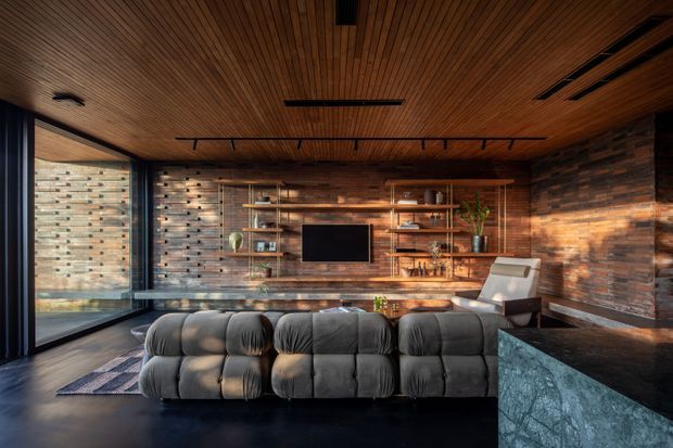 A warm, moody contemporary living room with a large modular charcoal sectional facing a wall-mounted television. The room features extensive dark timber lining on the ceiling, exposed brick walls, open timber shelving, black-framed floor-to-ceiling glazing, and a dark stone-clad foreground element, likely a kitchen or bar counter. Linear track lighting and integrated ceiling fixtures add a refined modern character.