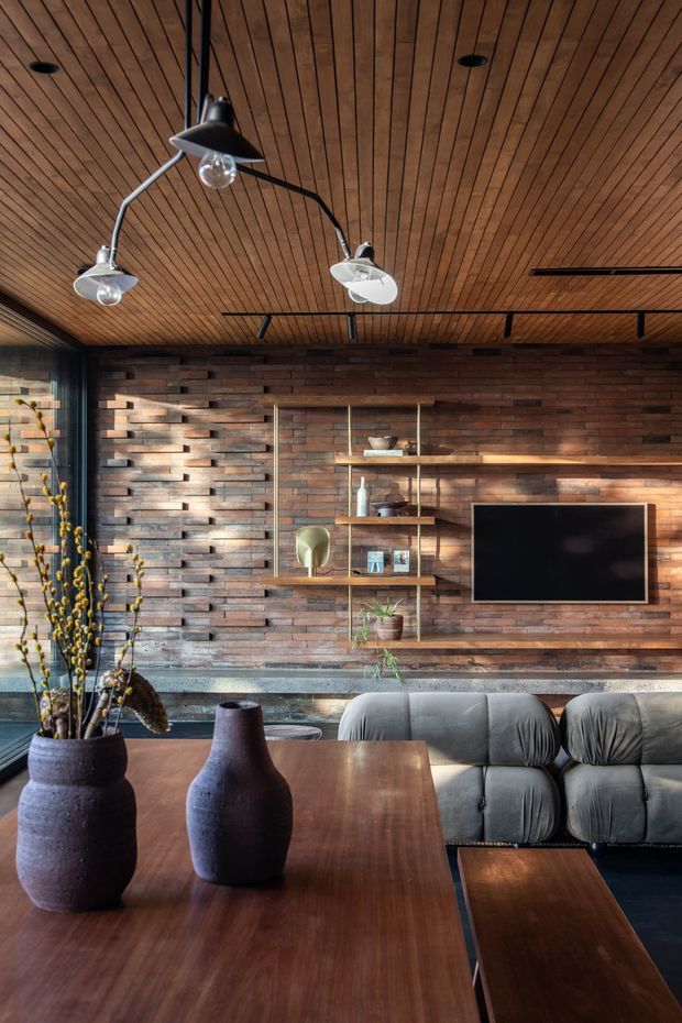 A warm, richly textured living room featuring a dark timber slatted ceiling, exposed dark brick feature wall, black-framed glazing, and a built-in timber shelving and television unit. A grey sectional sofa faces the wall-mounted TV, while a large timber coffee table with dark ceramic vases occupies the foreground. Sculptural black pendant lights and recessed spotlights add a refined, mid-century-inspired character.